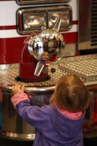 A toddler checks out her reflection in the fire truck's shiny surfaces.